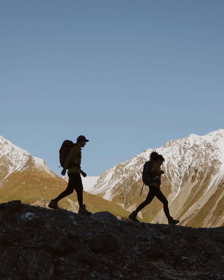 Two hikers crossing a ridge with snow-capped mountains behind